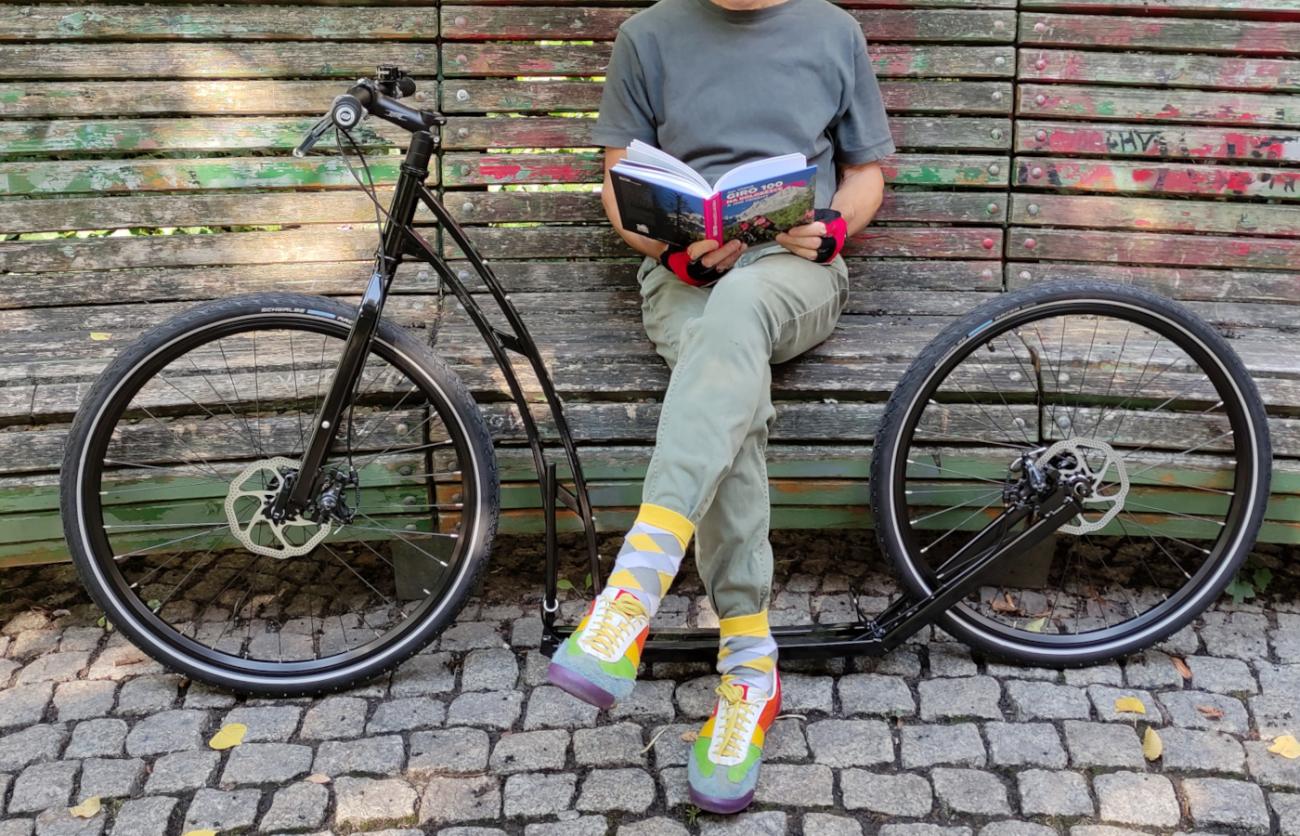 Lesend auf einer Parkbank, mit den Beinen zwischen den großen Laufrädern eines Tretrollers für Erwachsene
