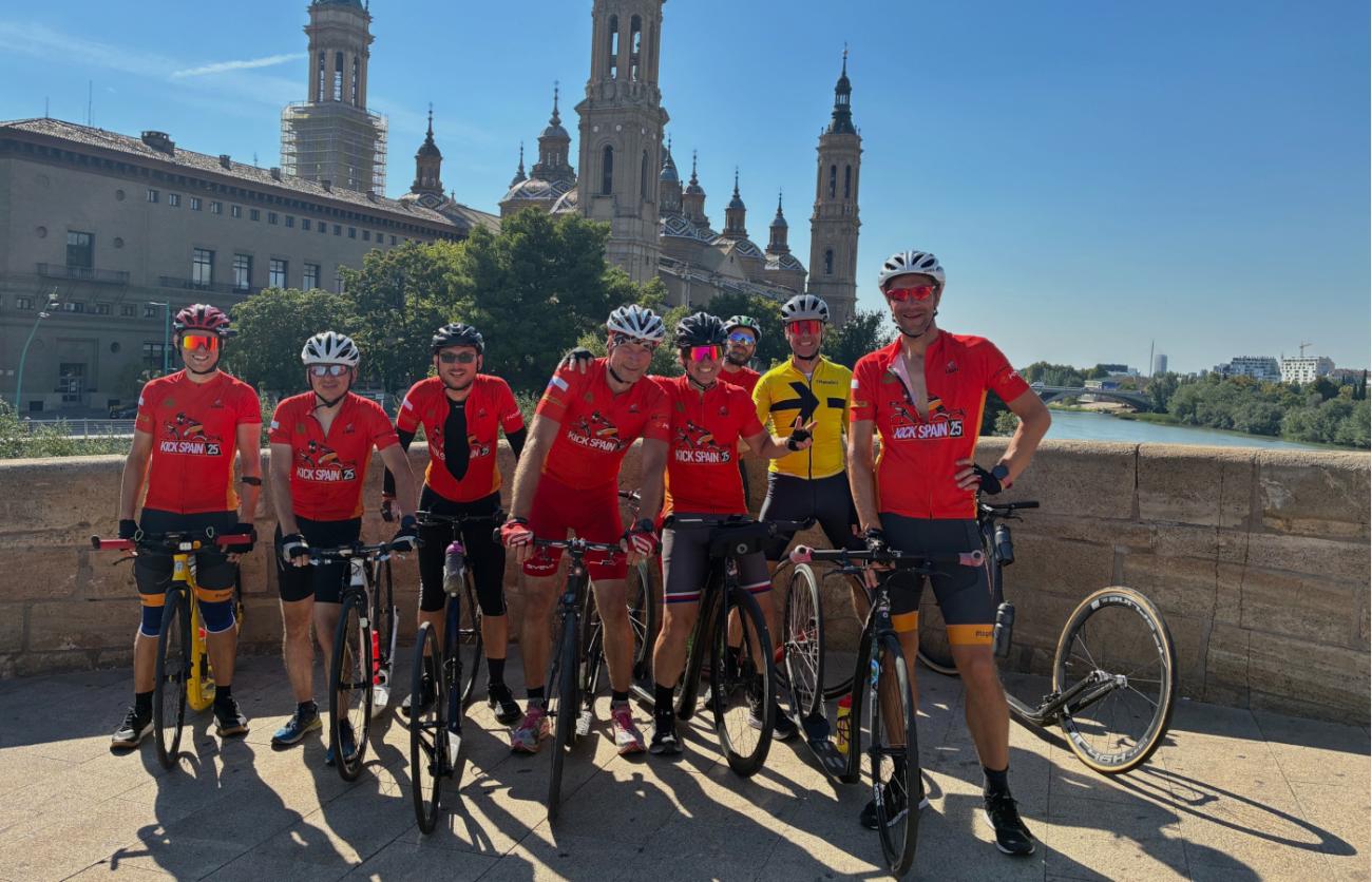 Acht Tretrollerfahrer in Trikots und Helmen vor der Kulisse einer spanischen Kirche.