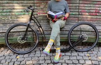 Lesend auf einer Parkbank, mit den Beinen zwischen den großen Laufrädern eines Tretrollers für Erwachsene